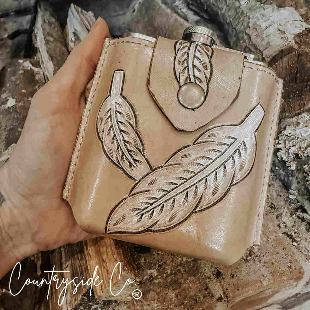 Hand holding a Desperado Tooled  Leather Flask with feather designs against a rustic background.