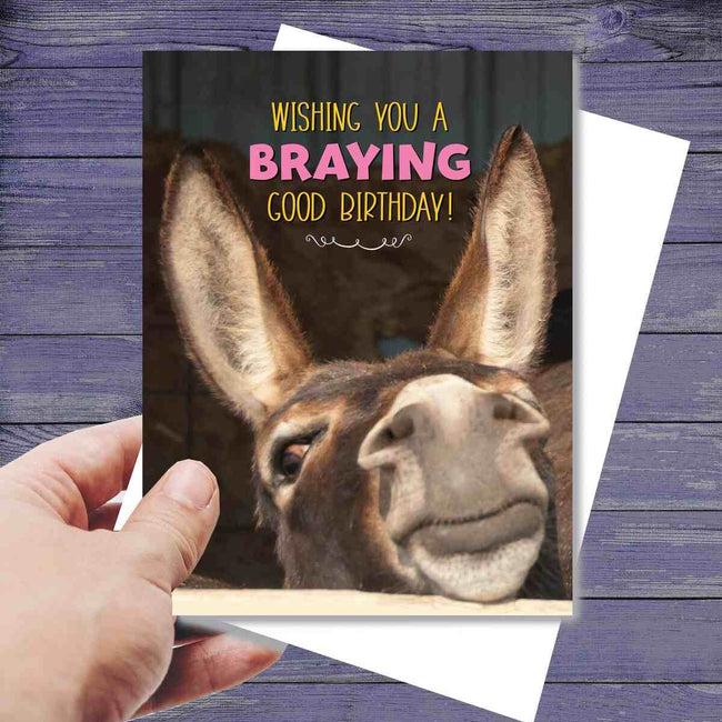 Hand holding a Donkey Birthday Card: Braying Good Birthday with a donkey's face and humorous text.