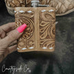 Hand holding a brown Leather Flask Stainless Steel with intricate patterns, against a dark background.