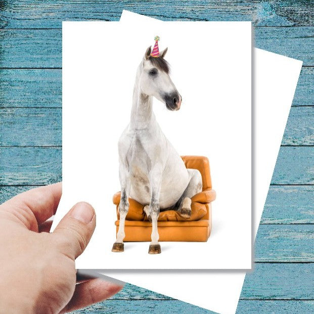 Horse Birthday Card: Horse Sitting - Horse on a couch with a birthday hat on a greeting card held by a hand against a blue wooden background.