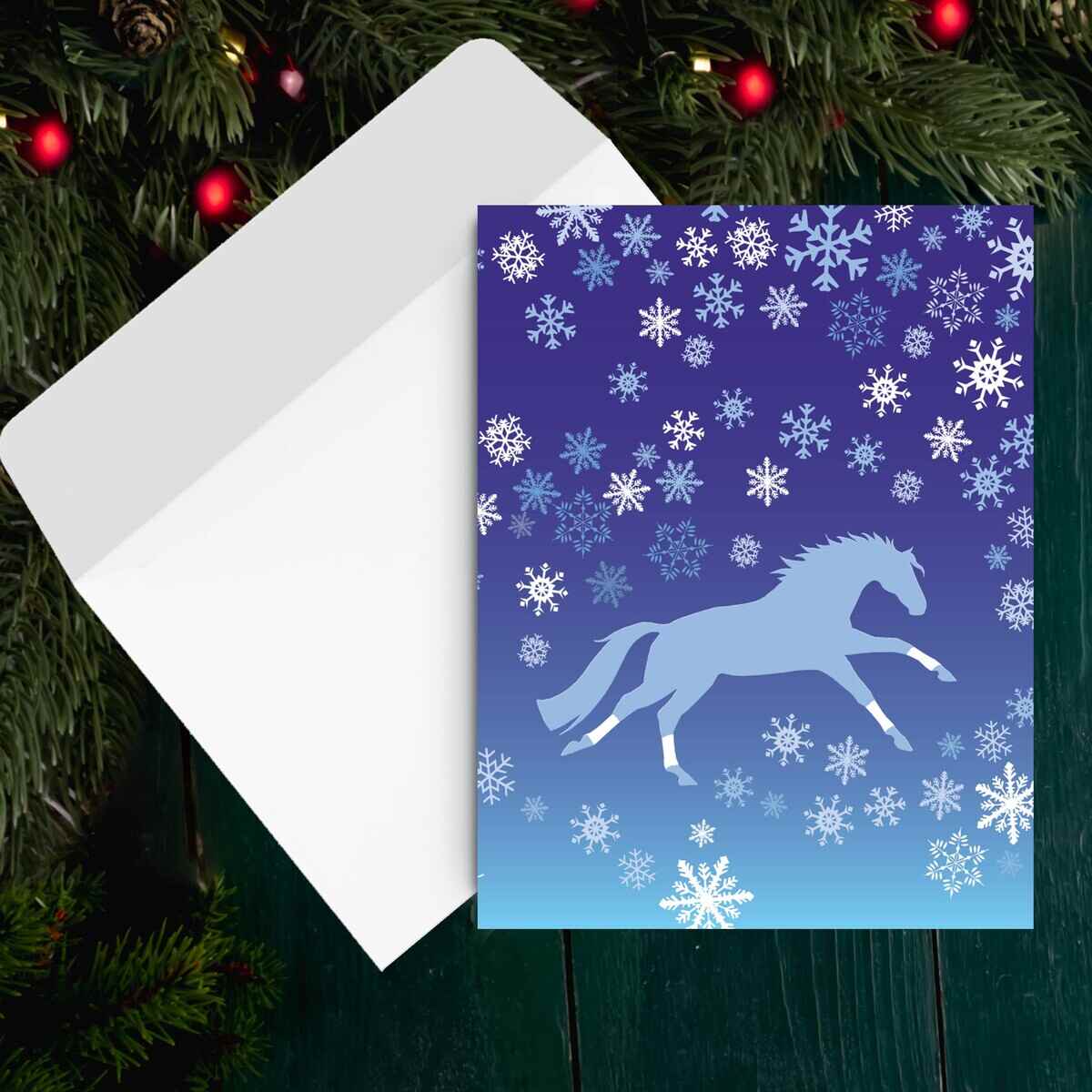 Horse Christmas Card - Galloping - with a blue horse design and snowflakes on a blue background, placed on a white envelope with a Christmas tree in the background.