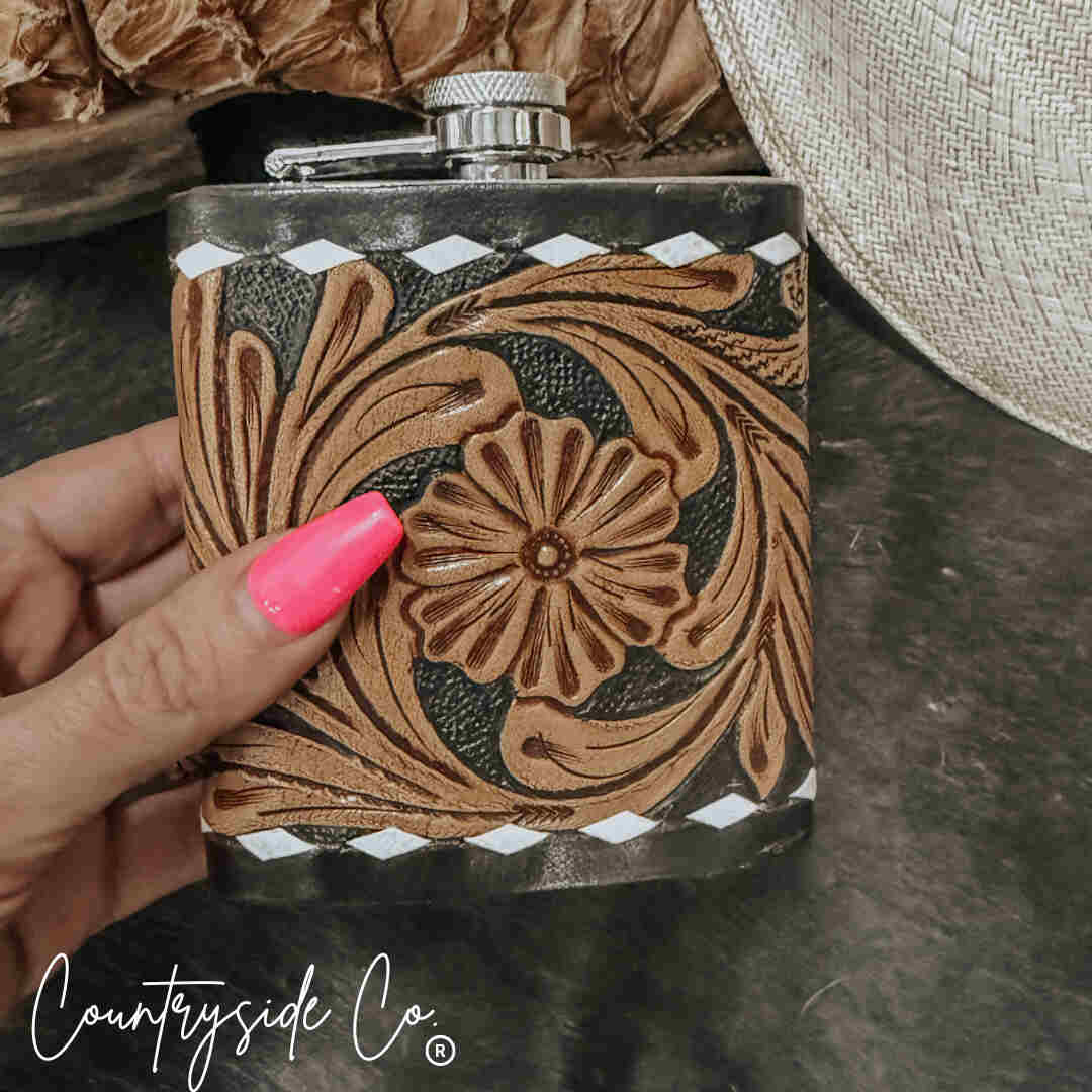 Hand holding a Leather Flask Stainless Steel with intricate brown designs on a dark surface, featuring 'Countryside Co' branding.