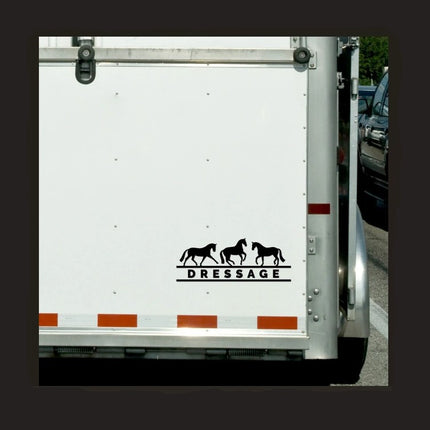 A vinyl decal with the word 'DRESSAGE' and silhouettes of horses above the text, placed on the side of a white horse trailer.